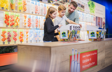 Ein Mann und zwei Kinder stehen am hölzernen Tresen des nachhaltigen Supermarktes der Klima Arena | © KLIMA ARENA Sinsheim/ Simon Hofmann