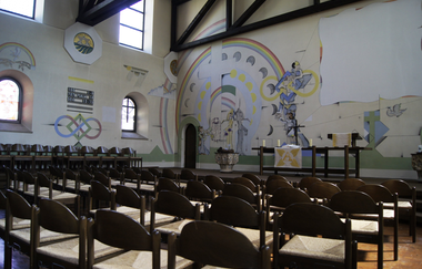 Bild zeigt die Altarwand in der Mauritiuskirche in Güglingen mit bunten Zeichnungen. Im Vordergrund stehen Stühle. | © Neckar-Zaber-Tourismus