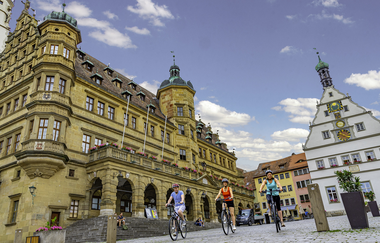 Blick auf den Marktplatz mit Rathaus in Rothenburg o.d.T.  Hier fahren gerade drei Radelnde über das Kopfsteinpflaster.