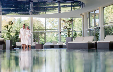 Im Innenraum der Therme spiegelt das Wasser die Fenster. Hinten steht ein Pärchen in Bademanteln. | © Solymar Therme