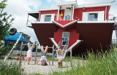 Blick auf Spielgeräte vor dem Toppels-Haus "Verdrehte Welt". Hier hängen drei Kinder mit dem Kopf nach unten an Kletterstangen. | © TOPPELS - Verdrehte Welt