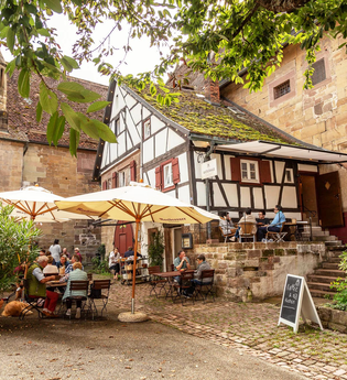 Wagnerhaus Cafe im Klosterhof Maulbronn mit Gästen an Tischen unter Sonnenschirmen, umgeben von historischen Gebäuden und Pflanzen. | © Wagnerhaus Café Maulbronn