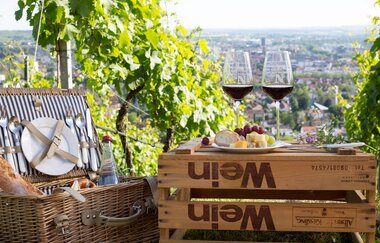 auf einer umgedrehten Weinkiste stehen zwei Gläser mit Rotwein und ein Teller mit Käse und Weintrauben daneben steht ein geflochtener Picknickkorb gefüllt mit Backwaren und Wasser am inneren des Deckels ist Besteck und Teller befestigt. | © Heilbronn Marketing GmbH | Maya Baum