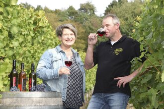 Eine Frau und ein Mann stehen in einem Weinberg bei einem Weinfass und halten ein Glas Rotwein in der Hand | © Touristikgemeinschaft Hohenlohe e. V. | Weinhof Dieroff