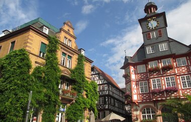 Links steht ein gelbes Sandsteingebäude mit grünem Bewuchs, rechts steht das Fachwerkbau des Rathauses mit großem Glockenturm. | © Maria Zimmermann