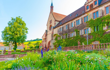 Blick aus dem blühenden Klostergarten auf das Kloster Bronnbach. Die Fassade ist begrünt. Im Hintergrund sind Steinfiguren und ein großer Baum zu sehen. | © Liebliches Taubertal