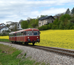 Zug mit zwei Waggons fährt auf Gleisen neben einem blühenden Feld und mehreren Häusern im Hintergrund | © Willi Hofmann