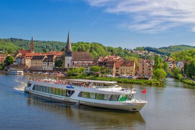 Fahrgastschiff auf dem Fluss vor der Stadt Neckargemünd mit historischen Gebäuden und bewaldeten Hügeln im Hintergrund. | © Stadt Neckargemünd, Foto Jens Hertel