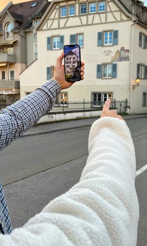 Zwei Personen zeigen mit ausgestrecktem Arm auf ein historisches Fachwerkhaus, das auf einem Smartphone-Bildschirm in einer Augmented-Reality-Ansicht zu sehen ist. | © Foto zeigmal GmbH