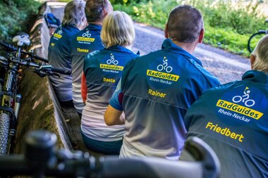 Gruppe von RadGuides Rhein-Neckar mit blauen Trikots und gelber Aufschrift sitzen auf einer Bank am Neckartalradweg mit Fahrrädern im Vordergrund | © Landratsamt Rhein-Neckar-Kreis; Foto Adrain Weng
