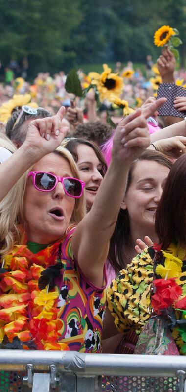 Menschenmenge bei einer Open-Air-Veranstaltung mit Sonnenblumen und bunten Blumenketten, einige heben die Arme | © Foto: Bernhardt Kreutzner
