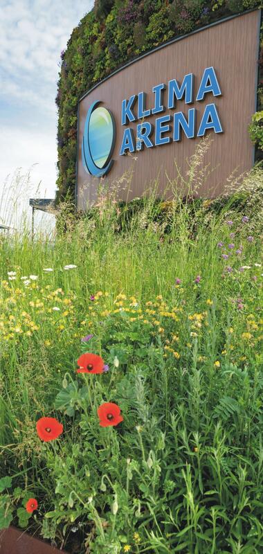 Holzfassade mit dem blauen Schriftzug 'KLIMA ARENA' und einem stilisierten Logo, davor eine Wiese mit Wildblumen und Mohnblumen. | © Landratsamt Rhein-Neckar-Kreis; Foto Dorothea Burkhardt