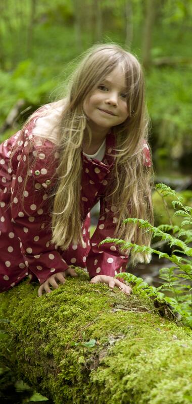 Kind mit langem Haar und gepunkteter Jacke sitzt auf moosbedecktem Baumstamm im Wald | © Landratsamt Rhein-Neckar-Kreis; Foto Dorothea Burkhardt