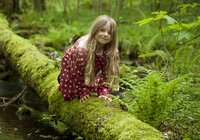 Kind mit langem Haar und gepunkteter Jacke sitzt auf moosbedecktem Baumstamm im Wald | © Landratsamt Rhein-Neckar-Kreis; Foto Dorothea Burkhardt