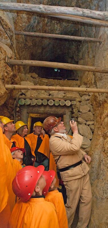 Gruppe von Personen in orangefarbener Schutzkleidung und roten Helmen in einem Stollen des Besucherbergwerks Schriesheim mit einem Führer, der auf eine Informationstafel an der Felswand zeigt. | © Landratsamt Rhein-Neckar-Kreis; Foto Dorothea Burkhardt