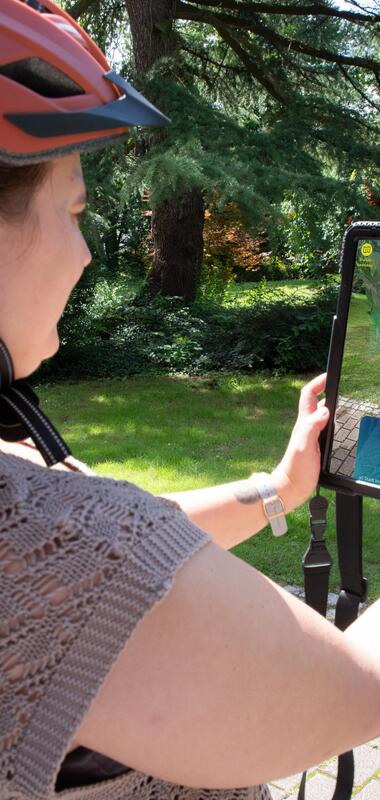 Frau mit orangefarbenem Helm hält Tablet, auf dem eine digitale Figur der Weitersehen App in einem Park angezeigt wird. | © Landratsamt Rhein-Neckar-Kreis; Foto Dorothea Burkhardt