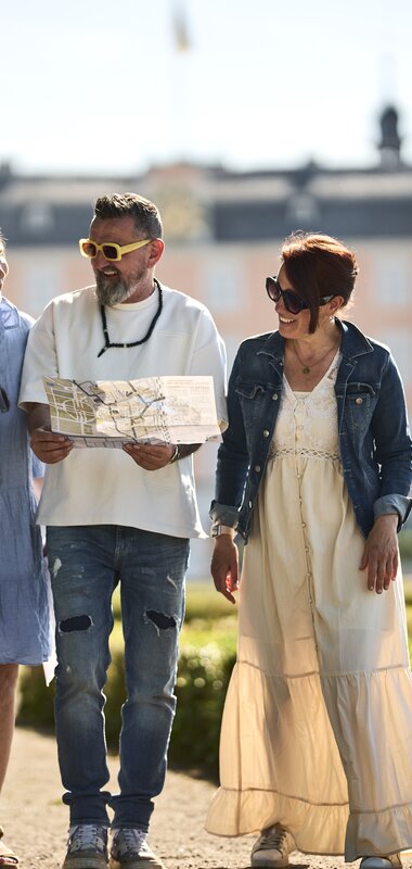 Drei Personen gehen auf einem Weg in einem Park vor dem Schwetzinger Schloss, eine Person hält eine Karte in der Hand. | © Rhein-Neckar-Kreis; Foto: Stefan Leitner