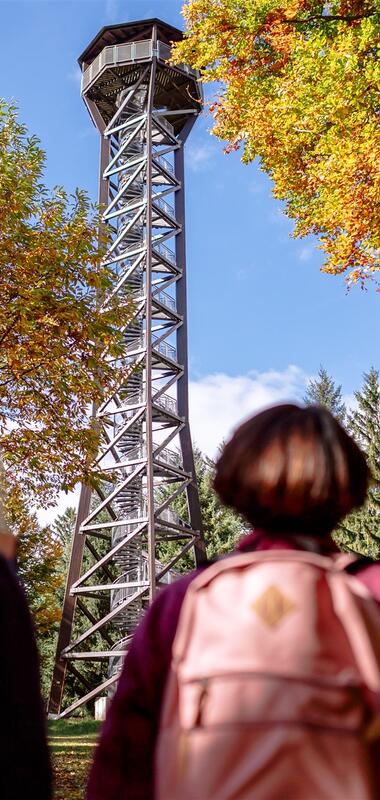 Zwei Personen mit Rucksäcken blicken auf den Teltschikturm, der von herbstlich gefärbten Bäumen umgeben ist | © Landratsamt Rhein-Neckar-Kreis; Foto Sebastian Weindel