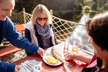 Personen sitzen an einem Holztisch im Freien an einem Gewässer, eine Person serviert einen Teller mit Fisch und Kartoffeln. | © Landratsamt Rhein-Neckar-Kreis; Foto Sebastian Weindel